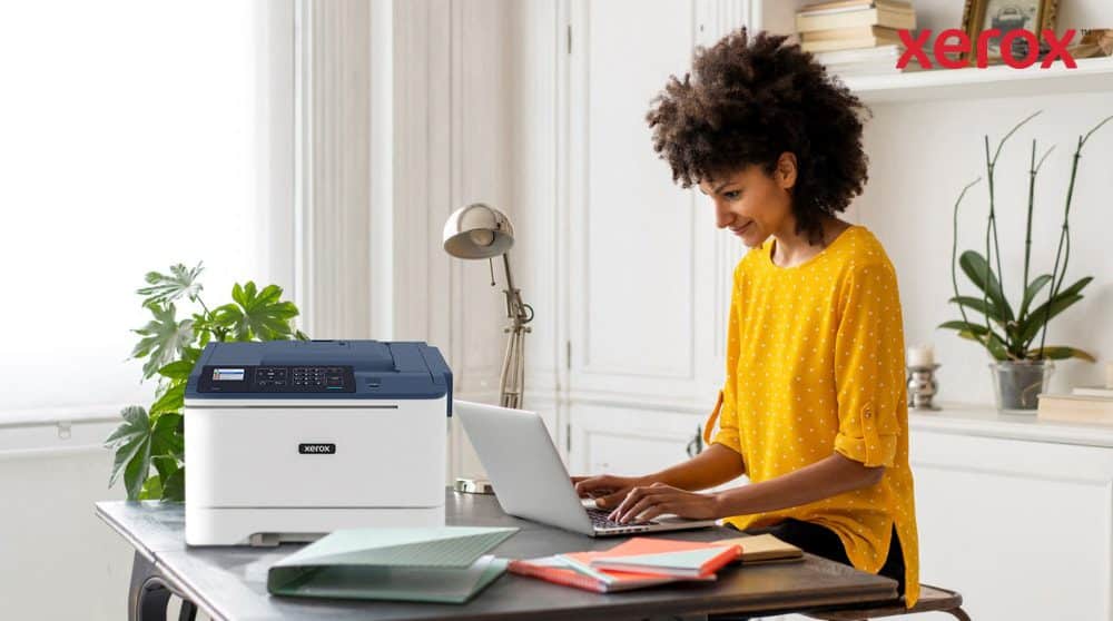 Xerox® DocuShare® Go: Produktivität für hybrides Arbeiten Eine Person mit lockigem Haar lächelt, während sie an einem Schreibtisch auf einem Laptop tippt. Neben dem Laptop steht ein kompakter Xerox C310-Drucker, der ein modernes Home-Office-Setup für mehr Produktivität vorführt.