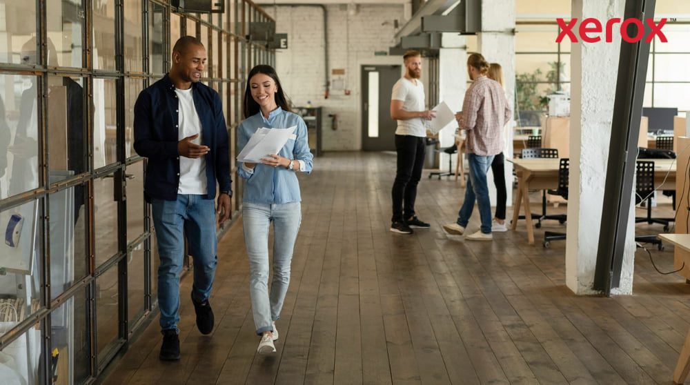 Xerox® Druck- und Scan-Erlebnis: Förderung der Zusammenarbeit am Arbeitsplatz Zwei Kollegen gehen lächelnd einen Flur in einem modernen Großraumbüro entlang, halten Dokumente in den Händen und sind im Hintergrund mit weiteren Kollegen zu sehen, was einen dynamischen und kollaborativen Arbeitsplatz darstellt.