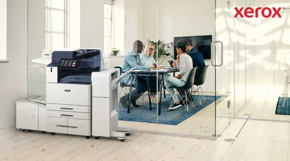 Xerox® Smart Start: Nahtlose Office-Integration Im Vordergrund eines hellen Büros steht ein moderner Multifunktionsdrucker Xerox AltaLink B8245. Im Hintergrund sitzt eine Gruppe von Menschen an einem Tisch und diskutiert gemeinsam.