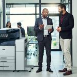Zwei Männer in einem Büro diskutieren etwas auf einem Tablet neben einem Farbmultifunktionsdrucker der Xerox® AltaLink® C8200-Serie Zwei Männer in einem Büro diskutieren etwas auf einem Tablet neben einem Farbmultifunktionsdrucker der Xerox® AltaLink® C8200-Serie